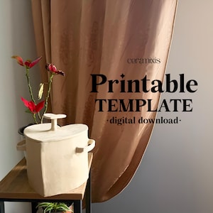 May include: A beige, heart-shaped ceramic container with a lid and handles, sitting on a wooden shelf. The image includes text that reads "Printable TEMPLATE digital download." Red and green plants are in the background.