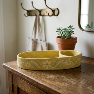 May include: Yellow ceramic oval tray with a floral design in white and light blue. The tray has a separate small compartment. A small potted plant sits nearby on a wooden surface. Decorative home decor.