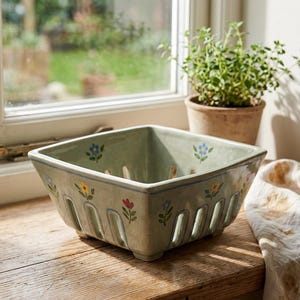 May include: A square, light green ceramic berry basket with floral designs in yellow, red, and blue. The basket has vertical slots for drainage and sits on a wooden surface. A small potted plant is visible in the background.