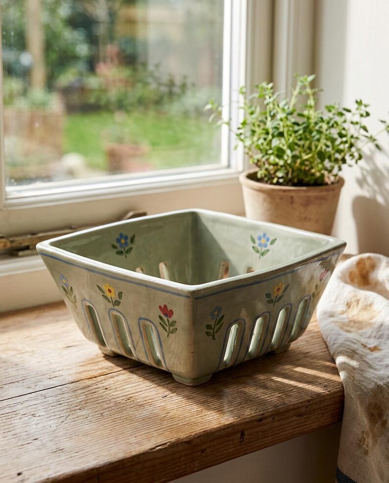 May include: A square, light green ceramic berry basket with floral designs in yellow, red, and blue. The basket has vertical slots for drainage and sits on a wooden surface. A small potted plant is visible in the background.