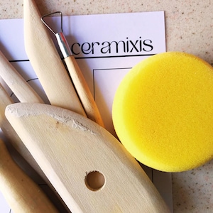 May include: A pottery tool kit with wooden tools, a metal and wood loop tool, and a yellow sponge. The tools are arranged on a white paper with the word "ceramixis" printed in black.