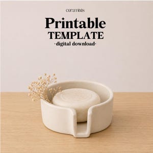 May include: A cream-colored ceramic soap dish with a round soap bar. The dish has a curved design with a small opening for drainage. The text "Printable TEMPLATE - digital download-" is at the top. A sprig of dried flowers adds a decorative touch.
