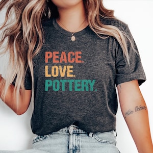 May include: Dark gray t-shirt with the words "PEACE LOVE POTTERY" in orange, yellow, and green. The shirt has a v-neck and short sleeves. The person is wearing light blue jeans and white sneakers.