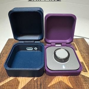 May include: Two small square boxes with lids, one is navy blue and the other is purple. The boxes are open and show the inside of the boxes. The navy blue box has a small grey circle in the centre of the bottom. The purple box has a silver circle with a black ring around it in the centre of the bottom. The boxes are sitting on a wooden surface with a star pattern.