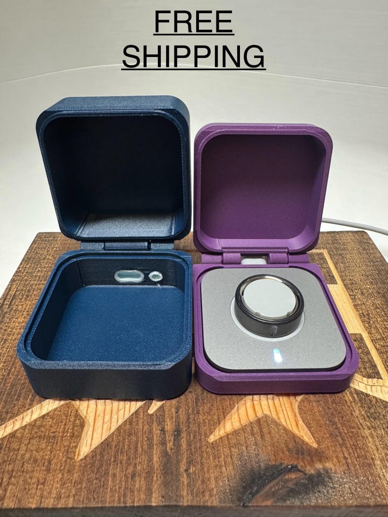 May include: Two small square boxes with lids, one is navy blue and the other is purple. The boxes are open and show the inside of the boxes. The navy blue box has a small grey circle in the centre of the bottom. The purple box has a silver circle with a black ring around it in the centre of the bottom. The boxes are sitting on a wooden surface with a star pattern.