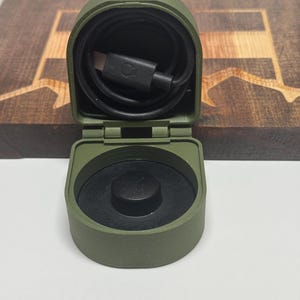 May include: A green, hinged, plastic case with a black charging cable inside. The case has a black button on the top.