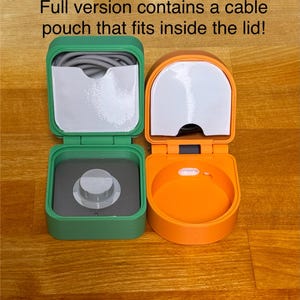 May include: Two small, square-shaped storage containers with lids. The container on the left is green and has a grey interior with a small knob. The container on the right is orange and has a grey interior. Both containers have a small, white, rectangular label on the lid.