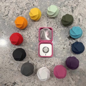 Puede incluir: Una colección de doce pequeños organizadores de cables de silicona con forma de espiral geométrica en varios colores, incluyendo rojo, naranja, amarillo, verde claro, verde oscuro, azul, marino, morado, rosa, blanco, gris y negro, rodeando un estuche cuadrado rosa.
