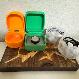 May include: Three small, 3D printed boxes, one orange, one green and one clear, are arranged on a wooden surface with a mountain design. The green box has a small, black disc inside.