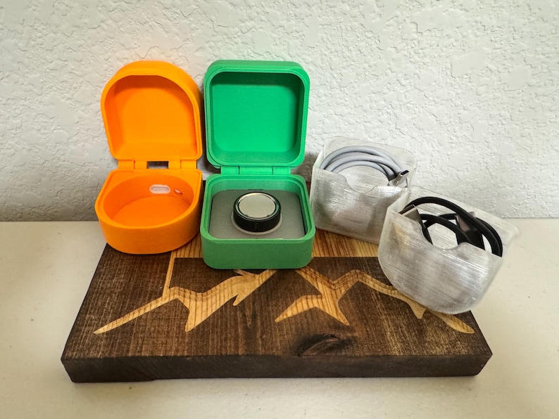 May include: Three small, 3D printed boxes, one orange, one green and one clear, are arranged on a wooden surface with a mountain design. The green box has a small, black disc inside.