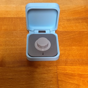May include: A small, light blue square box with a lid that is open. Inside the box is a small, grey knob with a black centre.