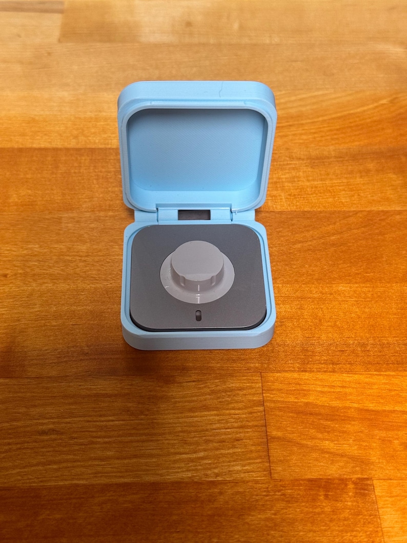 May include: A small, light blue square box with a lid that is open. Inside the box is a small, grey knob with a black centre.