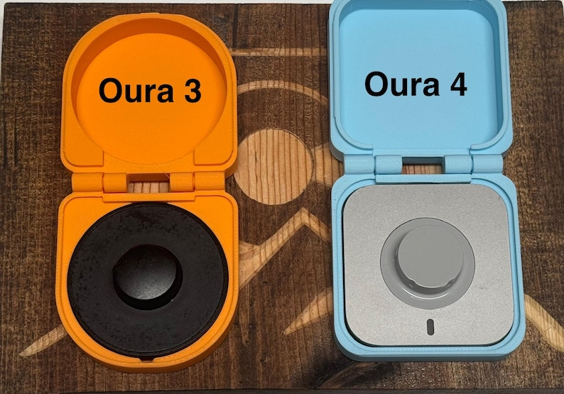 May include: Two small, round, electronic devices with lids. The device on the left is orange and says "Oura 3" on the lid. The device on the right is blue and says "Oura 4" on the lid.