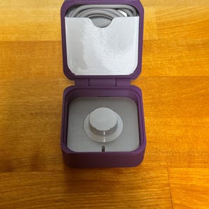 May include: A purple, square-shaped storage case with a white knob on the lid. The case is open and a grey cable is visible inside.