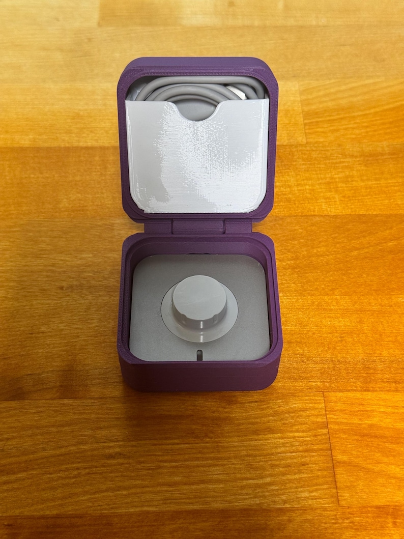May include: A purple, square-shaped storage case with a white knob on the lid. The case is open and a grey cable is visible inside.