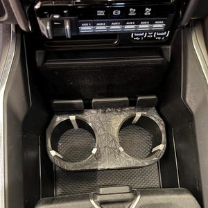 May include: A black plastic cup holder with two cup slots. The cup holder is designed to fit into a car's center console. The cup holder has a geometric pattern on the bottom.