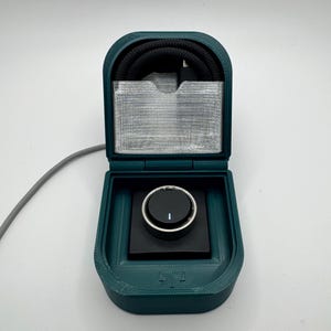 Puede incluir: Una caja de color verde azulado impresa en 3D con una perilla negra y un cable negro en su interior. La caja está abierta y la perilla es visible.