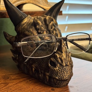May include: A detailed, bronze-colored dragon head sculpture with black horns, holding a pair of eyeglasses. The glasses have a silver frame and rectangular lenses. The dragon head is resting on a wooden surface.