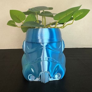 Puede incluir: Una maceta de plástico azul con forma de casco de Stormtrooper de Star Wars con una planta verde que crece en la parte superior.