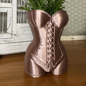 May include: A bronze-colored corset-shaped vase filled with white daisies. The vase has a detailed design with a lace-up front and a feminine silhouette. The daisies have white petals and yellow centers.
