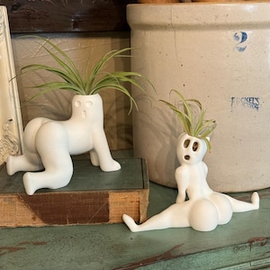 May include: Two white ceramic planters shaped like human figures, one crawling and the other reclining. Each planter holds a green plant with long, thin leaves. The planters are on a distressed teal surface.