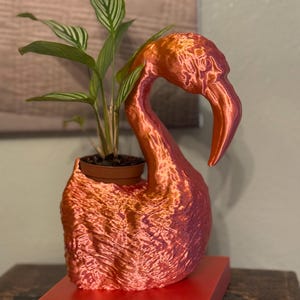 May include: A bright orange flamingo planter with a green plant inside. The planter is made of a textured material and has a base.