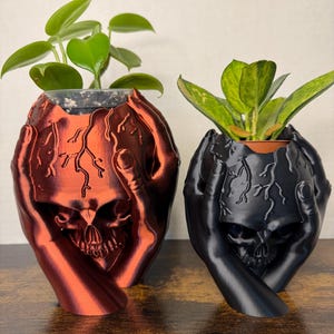 May include: Two skull planters, one in black and one in red, with hands holding the skull. Both planters have a plant growing out of the top.