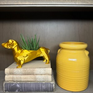 May include: A gold geometric dachshund figurine sits on a stack of books with a yellow ceramic jar on a wooden shelf. The dachshund is holding a small succulent plant.