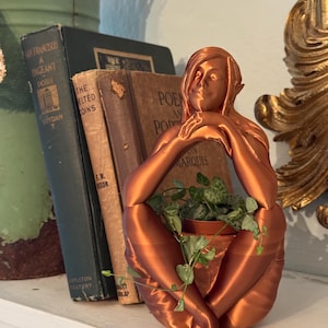 May include: A copper-colored planter shaped like a seated woman with a plant growing out of the top. The woman is sitting with her legs crossed and her hands resting on her chin. The plant is a trailing vine with green leaves.