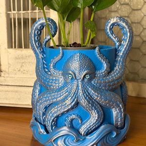 May include: A blue octopus-themed planter with silver accents and realistic detailing. The planter features an octopus design with tentacles and a detailed face. The planter holds a green leafy plant. The planter is a unique decorative item.