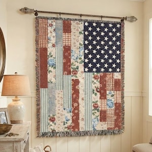May include: A woven wall tapestry featuring a patchwork design of the American flag, composed of vertical strips with floral, plaid, and striped patterns in red, white, and blue. The rectangular textile has a fringed edge and is hung on a metal rod.
