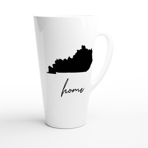 May include: A white ceramic latte mug featuring a black silhouette of the state of Kentucky. The word "home" is written in a cursive font below the state outline. The mug has a tall, tapered shape and a curved handle.