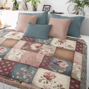 May include: A patchwork throw blanket with a floral pattern in shades of pink, blue, and brown, with a fringed edge. The blanket is on a bed with matching pillows in pink and teal. Decorative plants and a framed butterfly art piece are in the background.
