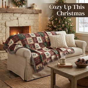 Könnte beinhalten: Ein beiges Sofa, bedeckt mit einer rot-grün-weißen karierten Decke mit Stechpalmenmustern und Fransen. Zwei cremefarbene Zopfstrickkissen liegen auf dem Sofa. Der Text "Cozy Up This Christmas" ist im oberen Bereich des Bildes zu sehen.