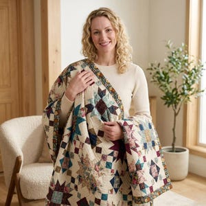 May include: A handmade quilt with a star pattern in cream, teal, burgundy, and navy. The quilt features a floral border and diamond shapes. The quilt is held by a person.