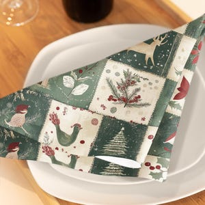 Puede incluir: Una servilleta plegada con temática navideña con un diseño de patchwork. La servilleta presenta ilustraciones de pájaros, ciervos, árboles de Navidad y bayas de acebo en tonos verdes, rojos y crema. La servilleta está colocada sobre un plato blanco.