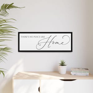 May include: A rectangular black framed wall art piece with the words "THERE'S NO PLACE LIKE Home" in black script. The artwork is displayed on a white wall above a light-colored wooden cabinet. A small potted plant and books are on the cabinet.