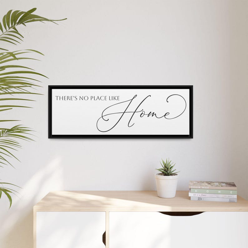 May include: A rectangular black framed wall art piece with the words "THERE'S NO PLACE LIKE Home" in black script. The artwork is displayed on a white wall above a light-colored wooden cabinet. A small potted plant and books are on the cabinet.