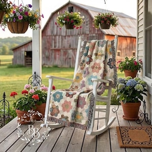 May include: A white wooden rocking chair draped with a woven throw blanket featuring a multi-colored floral hexagon pattern. The chair sits on a wooden porch surrounded by several terracotta pots filled with vibrant pink, red, and blue flowers.