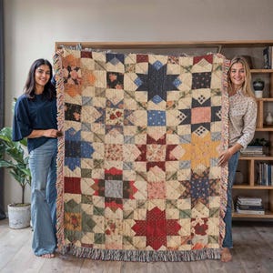 May include: A patchwork quilt featuring a star design in shades of red, blue, and yellow. The quilt has a fringed border and is displayed by two people. The quilt is constructed from a combination of squares and triangles.