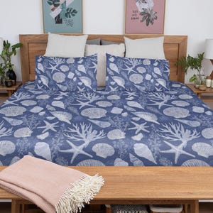 May include: A bed with a blue and white seashell pattern duvet cover and pillowcases. The bed is made up with a wooden headboard and a wooden bench at the foot of the bed. A pink blanket with fringe is on the bench.