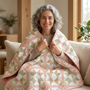 May include: A quilted blanket featuring a repeating geometric pattern of triangles in soft shades of sage green, dusty pink, and white. The quilt is constructed with a grid of pink and white gingham sashing and finished with a solid pink border.