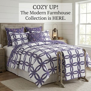 Può includere: Un letto con una trapunta bianca e federe abbinate con un motivo a cerchi viola intrecciati. Il testo COZY UP! The Modern Farmhouse Collection is HERE. è visualizzato sopra. Una lanterna in metallo si trova sul comodino in legno.