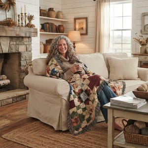 May include: A multi-colored patchwork throw blanket featuring a variety of geometric patterns in shades of red, blue, green, and tan. The blanket has a fringed edge and is draped over a person sitting on a beige sofa in a living room setting.