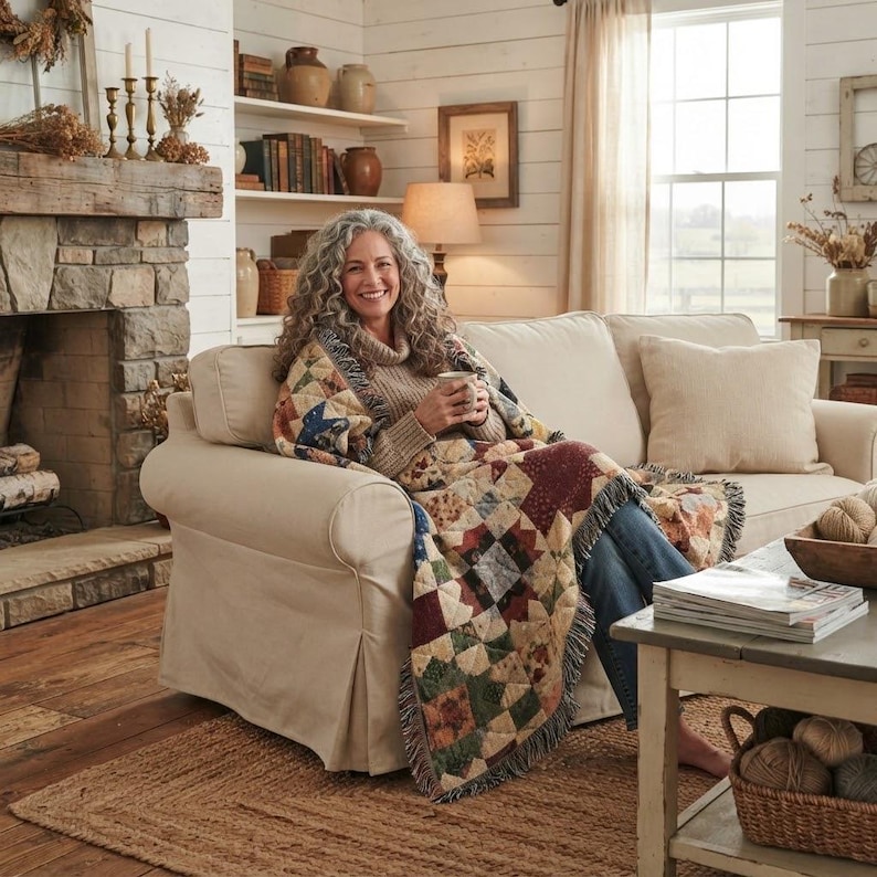 May include: A multi-colored patchwork throw blanket featuring a variety of geometric patterns in shades of red, blue, green, and tan. The blanket has a fringed edge and is draped over a person sitting on a beige sofa in a living room setting.