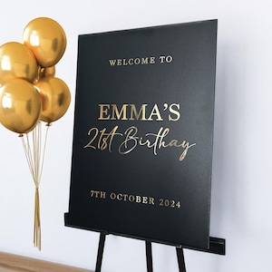 May include: A black sign on a black easel with gold lettering that reads "WELCOME TO EMMA'S 21st Birthday 7th October 2024". A cluster of gold balloons is tied to the left of the sign.