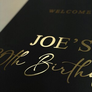 May include: Black card with gold foil lettering that reads "WELCOME" and "JOE'S 70th Birthday". The text is in a mix of serif and script fonts. The card is angled, showing a close-up of the text.
