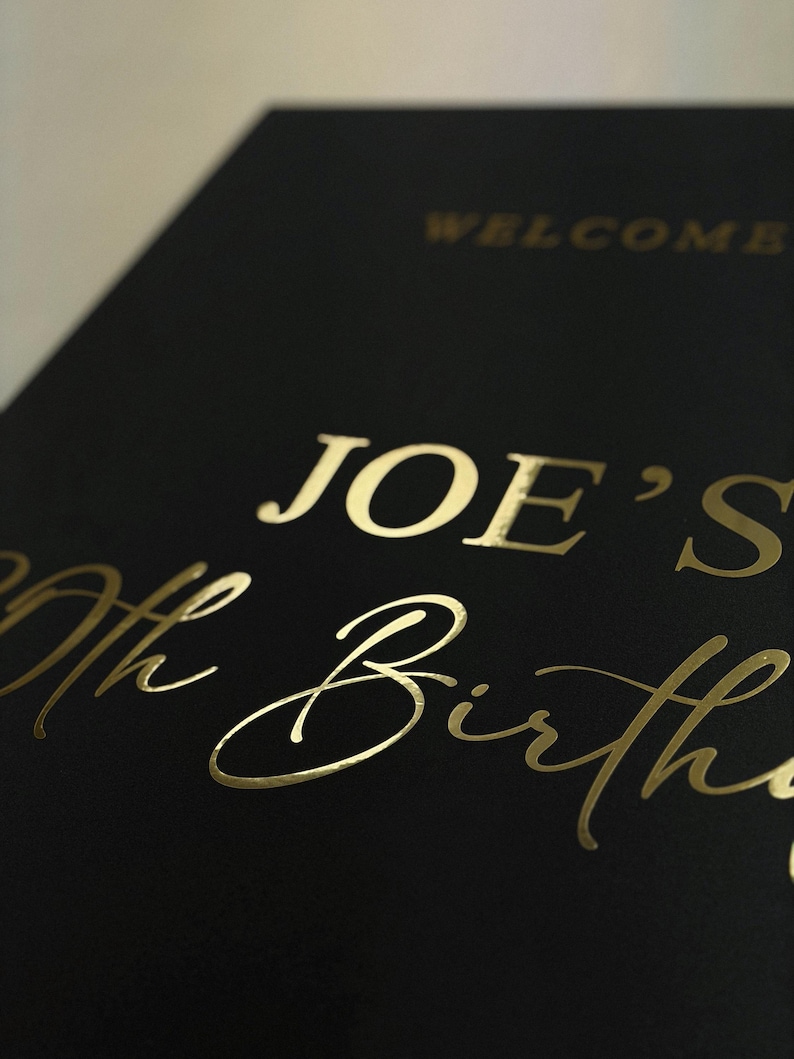 May include: Black card with gold foil lettering that reads "WELCOME" and "JOE'S 70th Birthday". The text is in a mix of serif and script fonts. The card is angled, showing a close-up of the text.