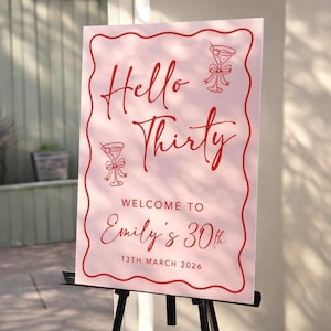 May include: A pink sign with red text that reads "Hello Thirty" and "Welcome to Emily's 30th". The sign includes illustrations of martini glasses and is displayed on a black easel. The date is March 13th, 2026.