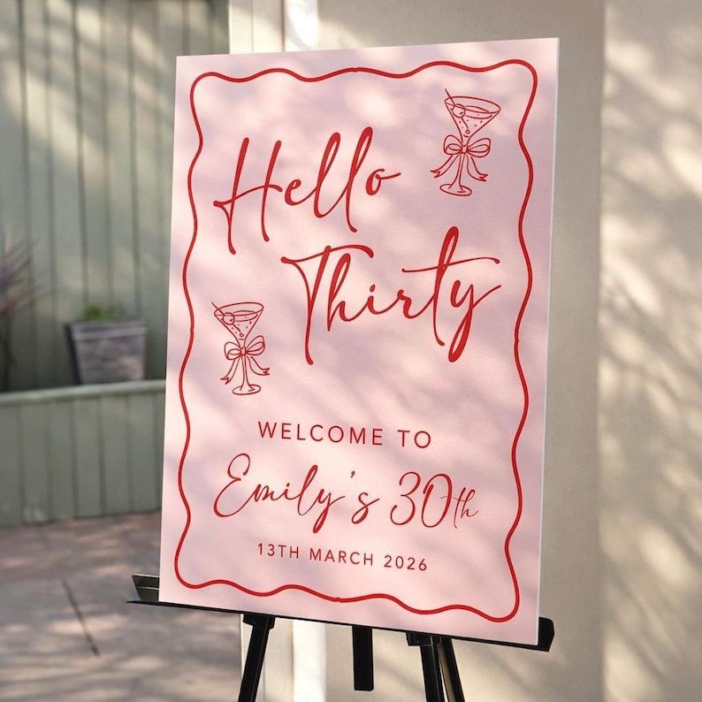 May include: A pink sign with red text that reads "Hello Thirty" and "Welcome to Emily's 30th". The sign includes illustrations of martini glasses and is displayed on a black easel. The date is March 13th, 2026.